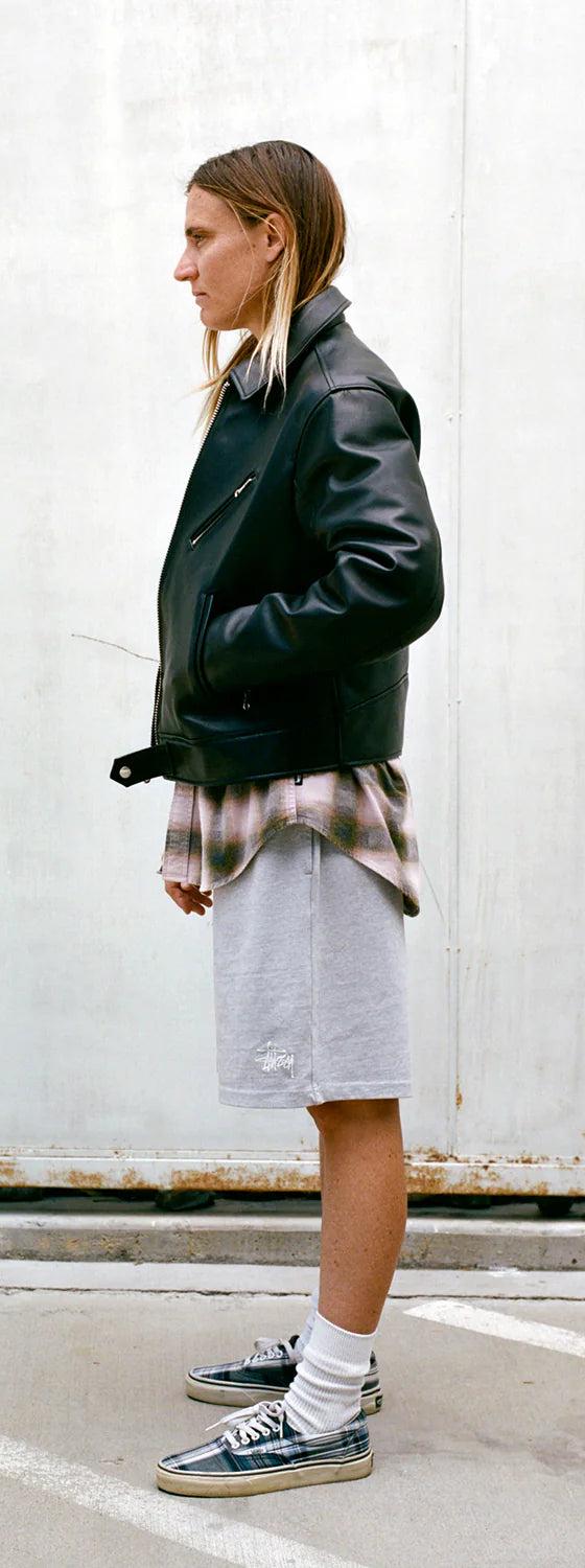 A person wearing a black leather jacket over a faded plaid shirt with grey Stüssy sweat shorts and sneakers