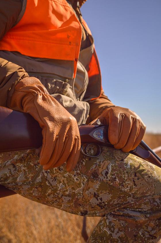 A hunter wearing tan Sullivan Glove Co leather gloves resting on a shotgun in an open field