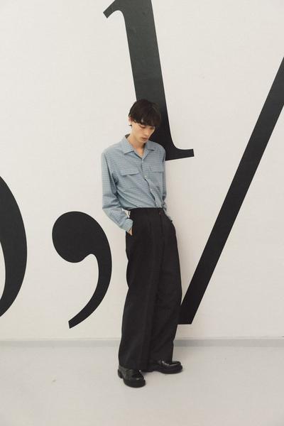 A man wearing a blue checked Sunflower overshirt with black wide-leg trousers