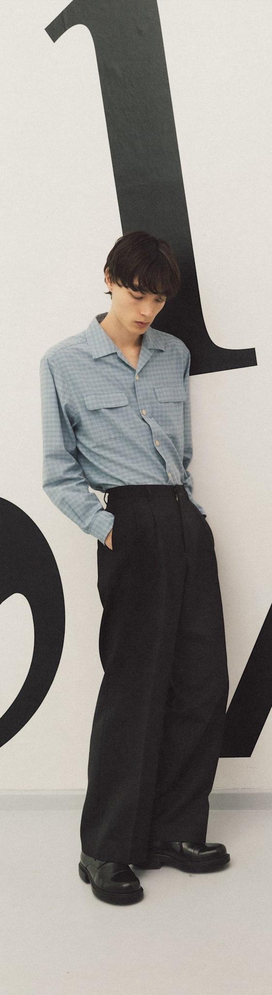 A man wearing a blue checked Sunflower overshirt with black wide-leg trousers