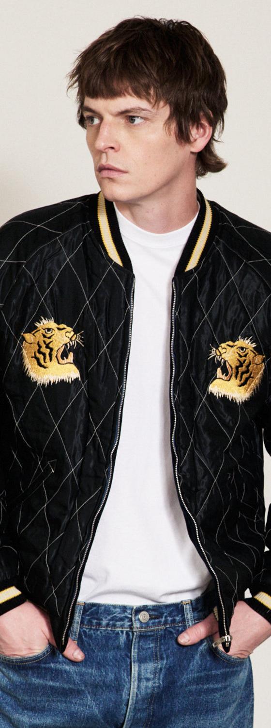 A man wearing a black quilted Tailor Toyo souvenir jacket with embroidered tiger heads over a white tee