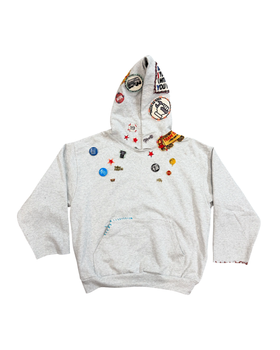 A grey Taller de Rafa hoodie covered in enamel pins and patches on a plain white background