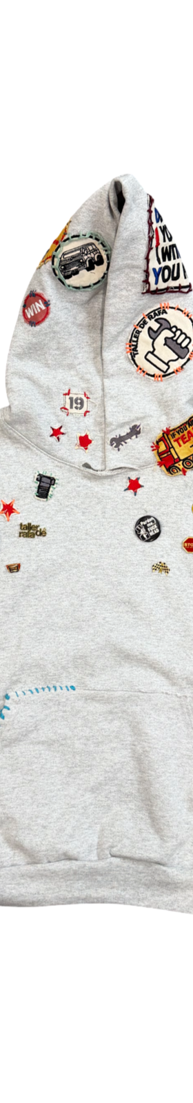A grey Taller de Rafa hoodie covered in enamel pins and patches on a plain white background