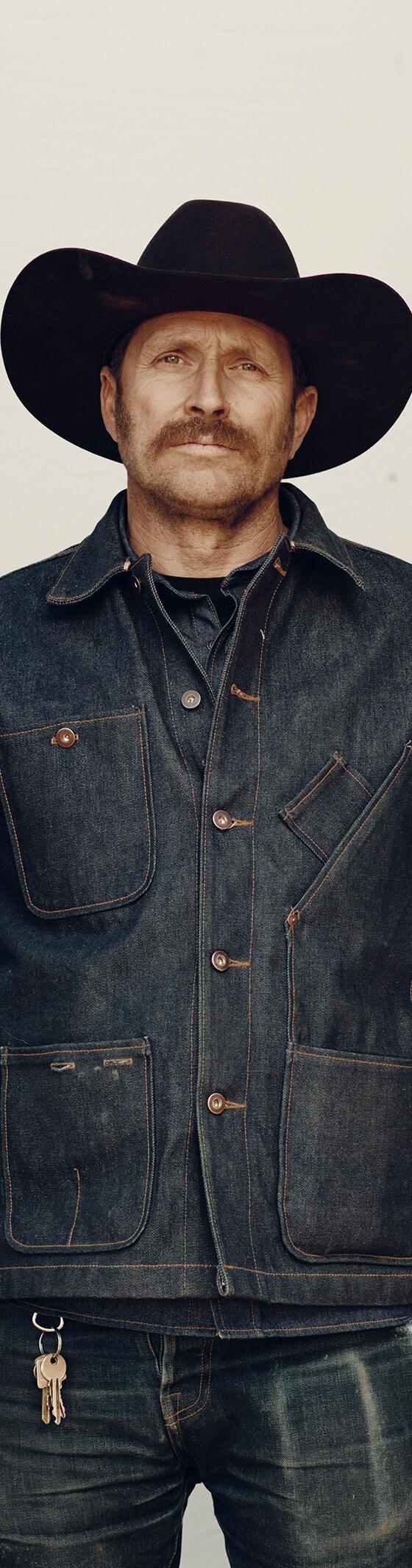 A man in a black cowboy hat wearing a raw indigo Tellason denim chore jacket and jeans