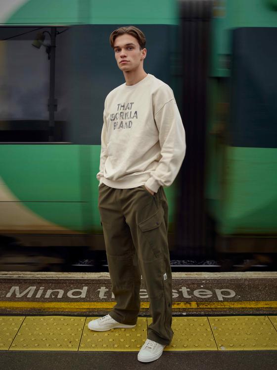 A man wearing a cream That Gorilla Brand sweatshirt and olive cargo trousers on a train platform