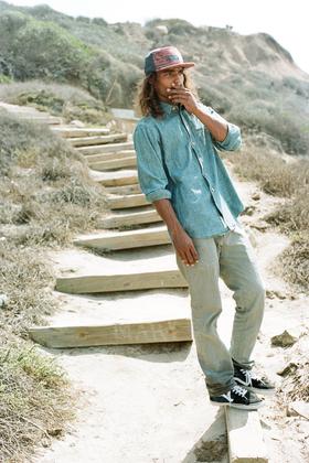 A man wearing a patterned five-panel The Ampal Creative cap with a chambray shirt and faded jeans on coastal steps