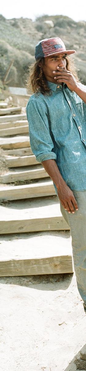 A man wearing a patterned five-panel The Ampal Creative cap with a chambray shirt and faded jeans on coastal steps