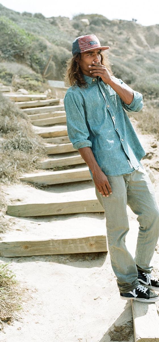 A man wearing a patterned five-panel The Ampal Creative cap with a chambray shirt and faded jeans on coastal steps