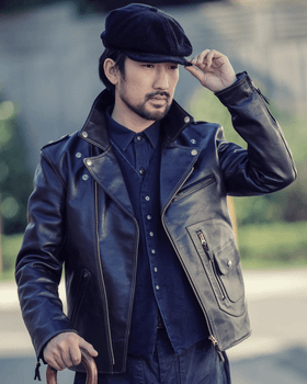 A man wearing a black The Flat Head horsehide leather jacket over an indigo denim shirt with a wool newsboy cap