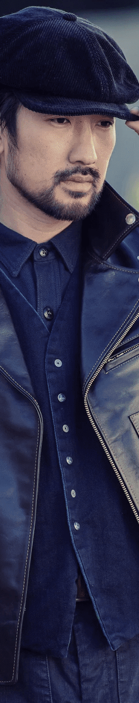 A man wearing a black The Flat Head horsehide leather jacket over an indigo denim shirt with a wool newsboy cap