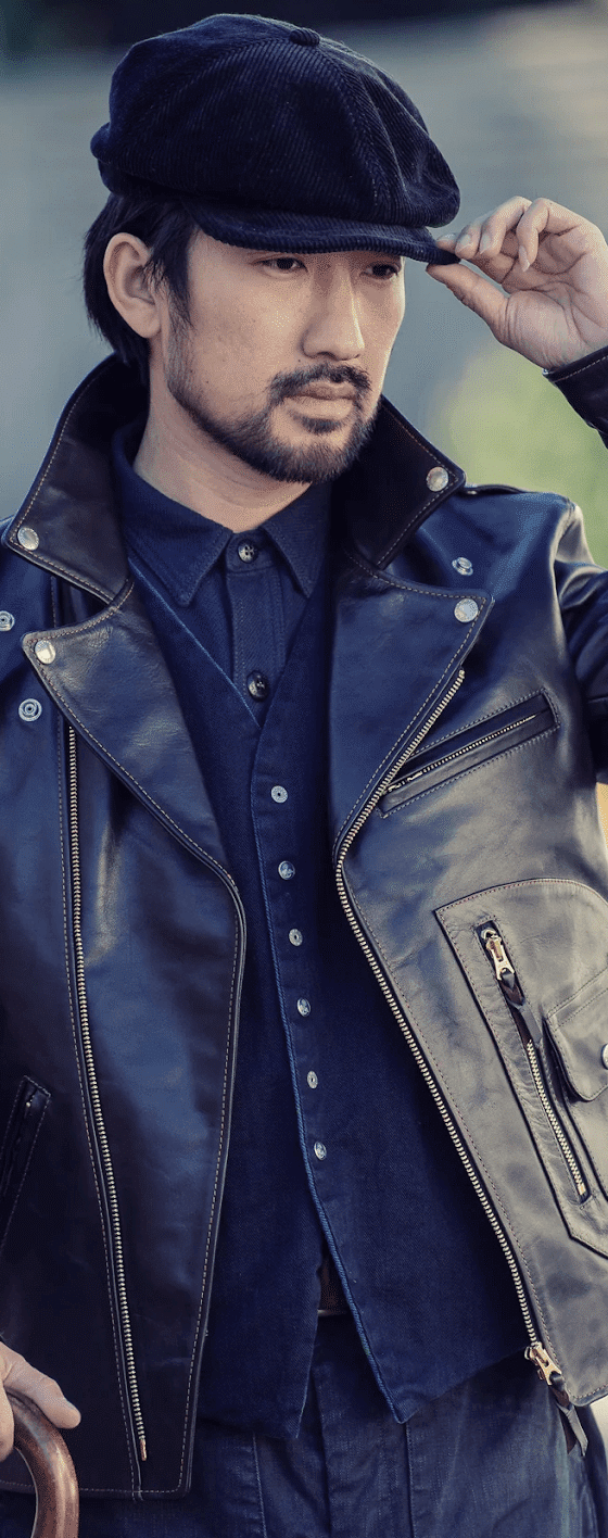 A man wearing a black The Flat Head horsehide leather jacket over an indigo denim shirt with a wool newsboy cap