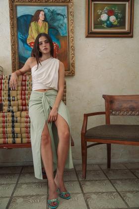 A woman wearing a one-shoulder draped cream top and sage green wrap skirt by The Line by K
