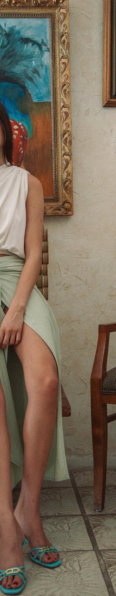 A woman wearing a one-shoulder draped cream top and sage green wrap skirt by The Line by K