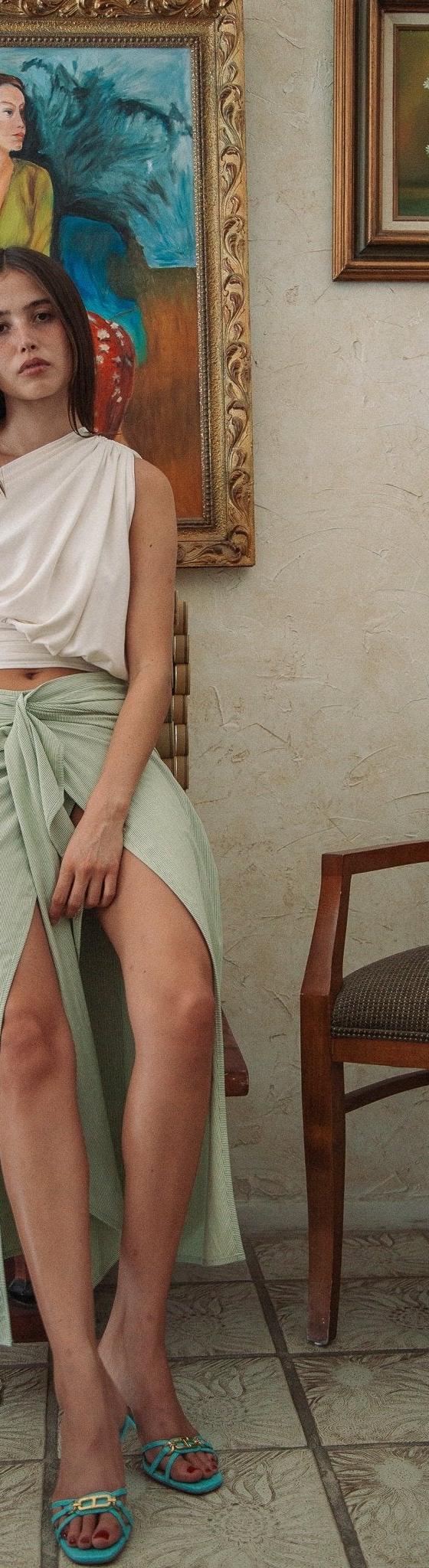 A woman wearing a one-shoulder draped cream top and sage green wrap skirt by The Line by K
