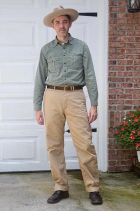 A man wearing a green chambray work shirt and tan high-waisted trousers with a wide-brim hat