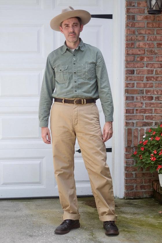 A man wearing a green chambray work shirt and tan high-waisted trousers with a wide-brim hat