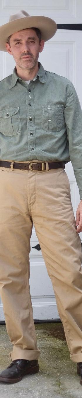 A man wearing a green chambray work shirt and tan high-waisted trousers with a wide-brim hat