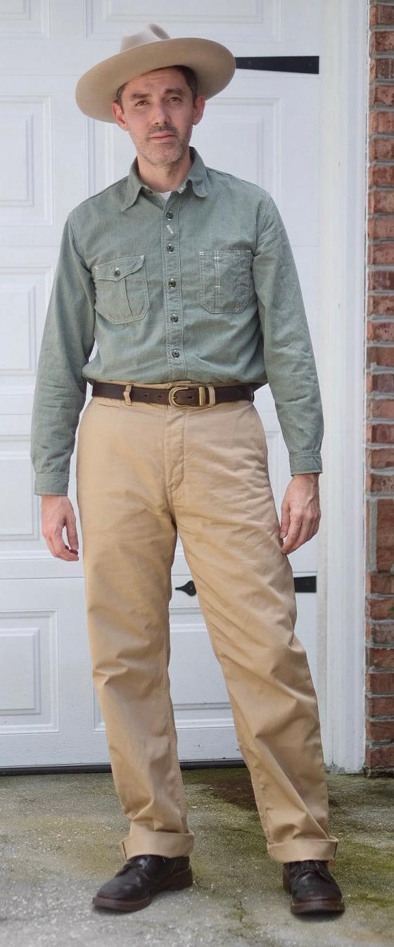 A man wearing a green chambray work shirt and tan high-waisted trousers with a wide-brim hat