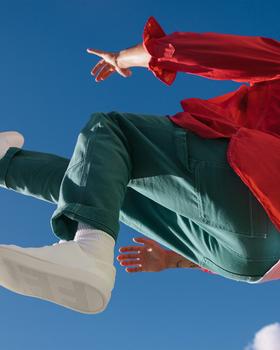 A person in a red jacket and green trousers leaping against a blue sky in white Thousand Fell low-top sneakers