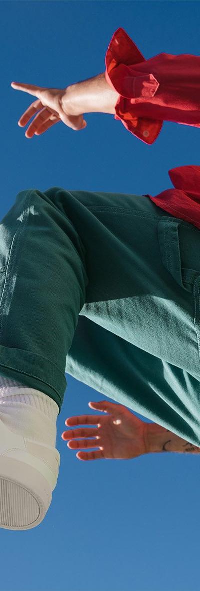 A person in a red jacket and green trousers leaping against a blue sky in white Thousand Fell low-top sneakers
