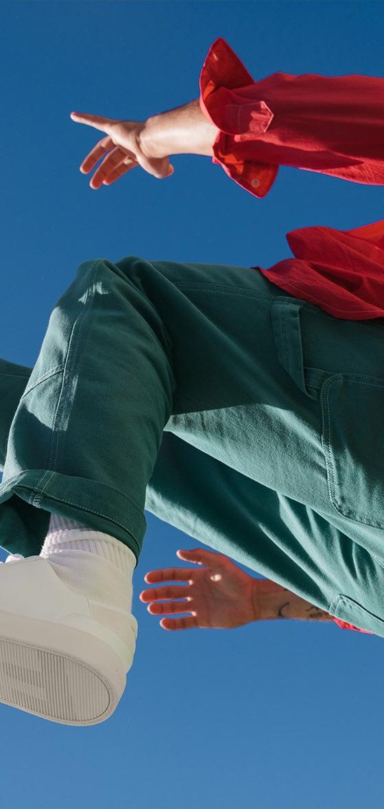 A person in a red jacket and green trousers leaping against a blue sky in white Thousand Fell low-top sneakers