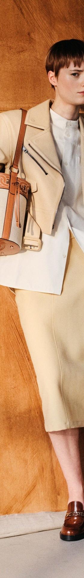 A woman wearing a cream Tod's leather jacket and knit skirt with a tan bucket bag and brown leather loafers