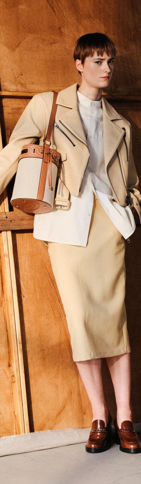 A woman wearing a cream Tod's leather jacket and knit skirt with a tan bucket bag and brown leather loafers