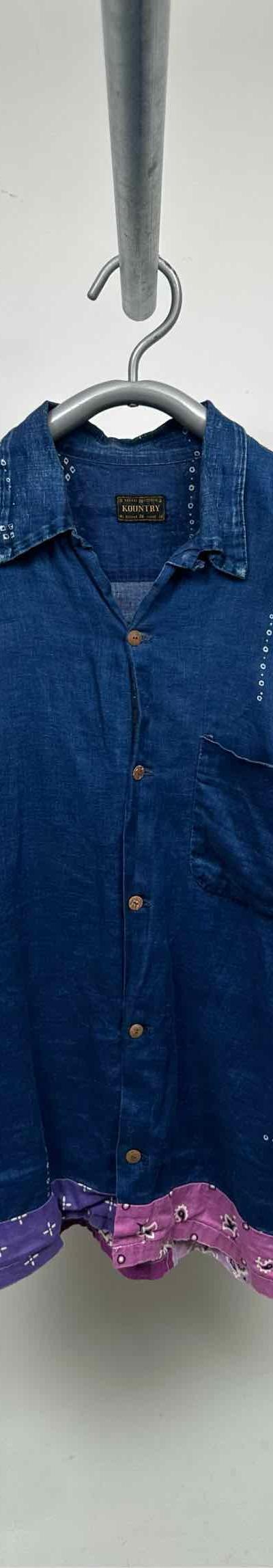 An indigo denim short-sleeve camp collar shirt with pink bandana patchwork hem, photographed on a hanger against a grey wall
