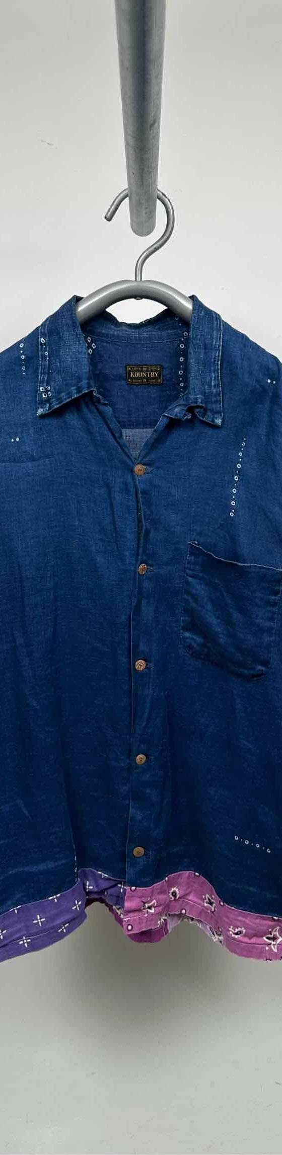 An indigo denim short-sleeve camp collar shirt with pink bandana patchwork hem, photographed on a hanger against a grey wall
