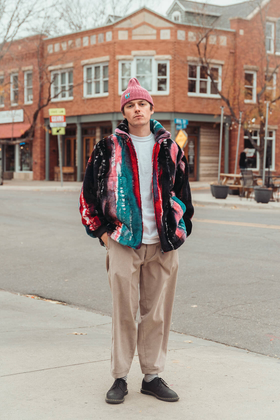 A man wearing a multicolour striped ts(s) fleece jacket with pleated beige trousers and a pink beanie