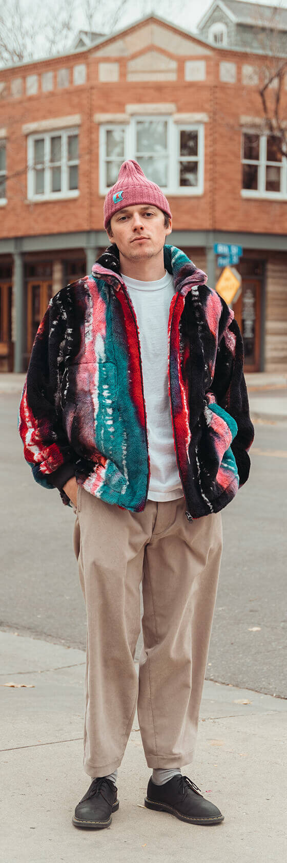 A man wearing a multicolour striped ts(s) fleece jacket with pleated beige trousers and a pink beanie