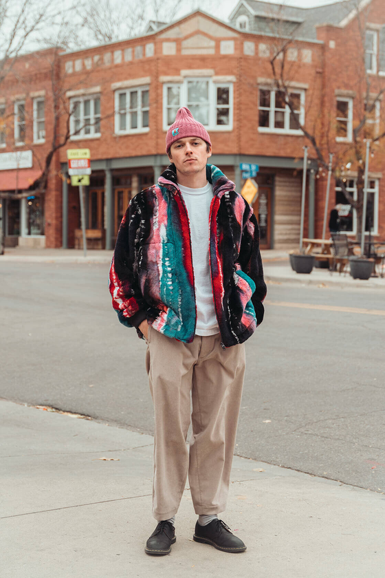 A man wearing a multicolour striped ts(s) fleece jacket with pleated beige trousers and a pink beanie
