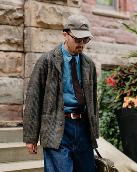 A man wearing a brown checked tweed chore jacket over a denim shirt and tie with wide indigo jeans