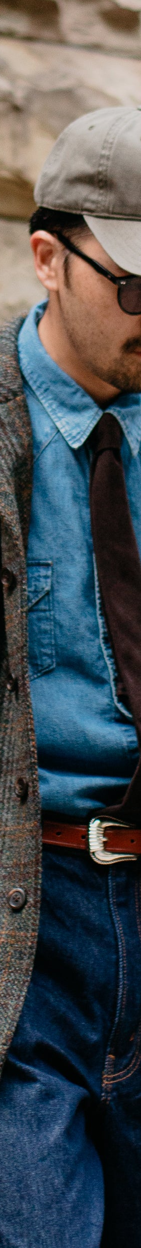 A man wearing a brown checked tweed chore jacket over a denim shirt and tie with wide indigo jeans