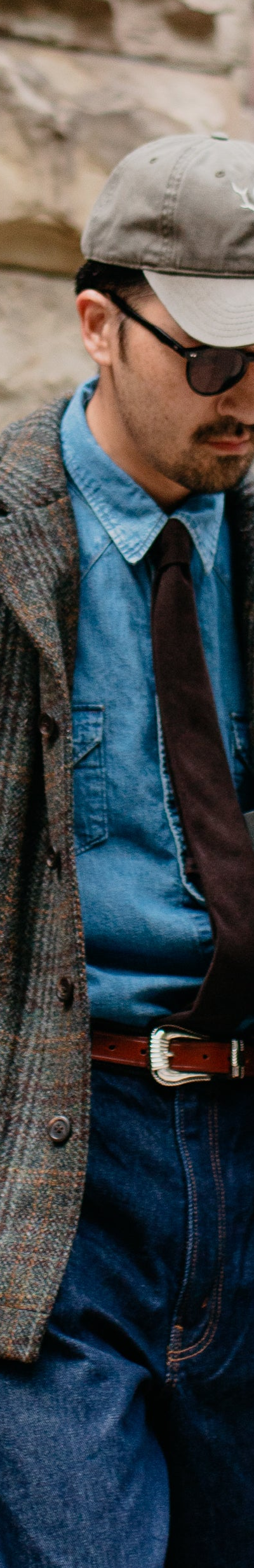 A man wearing a brown checked tweed chore jacket over a denim shirt and tie with wide indigo jeans
