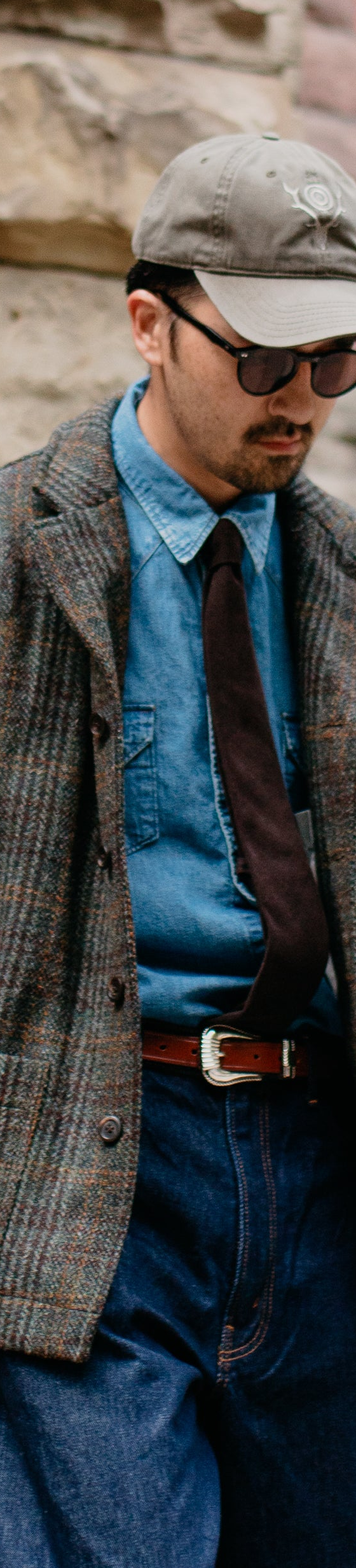 A man wearing a brown checked tweed chore jacket over a denim shirt and tie with wide indigo jeans