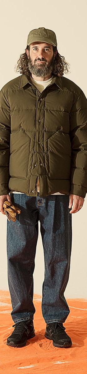 A man wearing an olive Uniform Bridge down jacket with a matching cap and dark denim jeans