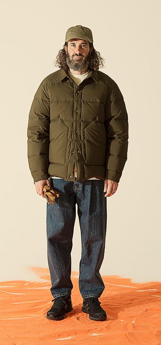 A man wearing an olive Uniform Bridge down jacket with a matching cap and dark denim jeans