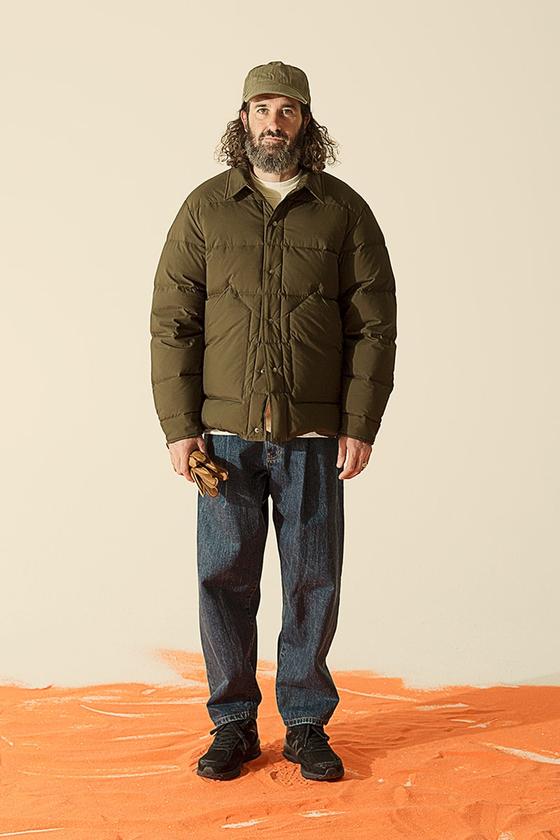 A man wearing an olive Uniform Bridge down jacket with a matching cap and dark denim jeans