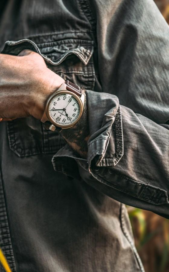 A man in a washed grey denim jacket wearing a Vortic Watch Company wristwatch with a white vintage dial and leather strap
