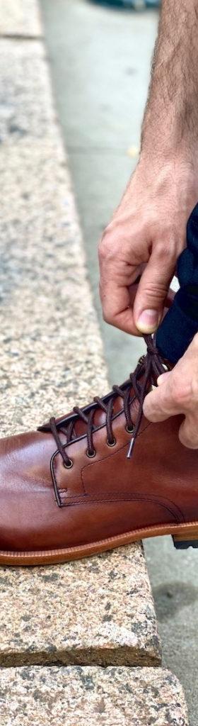 A man lacing a brown Warfield &amp; Grand leather boot on a curb
