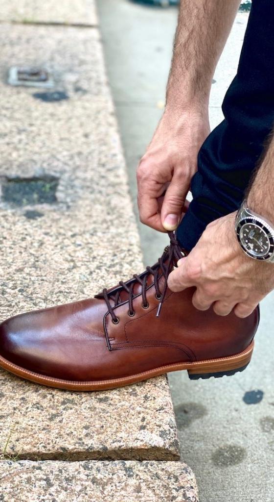 A man lacing a brown Warfield &amp; Grand leather boot on a curb