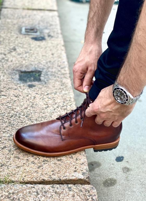 A man lacing a brown Warfield & Grand leather boot on a curb