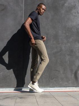 A man wearing olive Western Rise technical travel pants with a black tee, leaning against a concrete wall