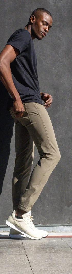 A man wearing olive Western Rise technical travel pants with a black tee, leaning against a concrete wall