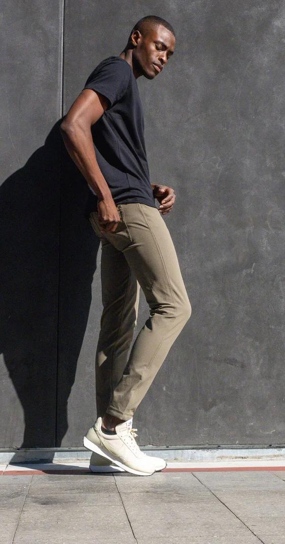 A man wearing olive Western Rise technical travel pants with a black tee, leaning against a concrete wall
