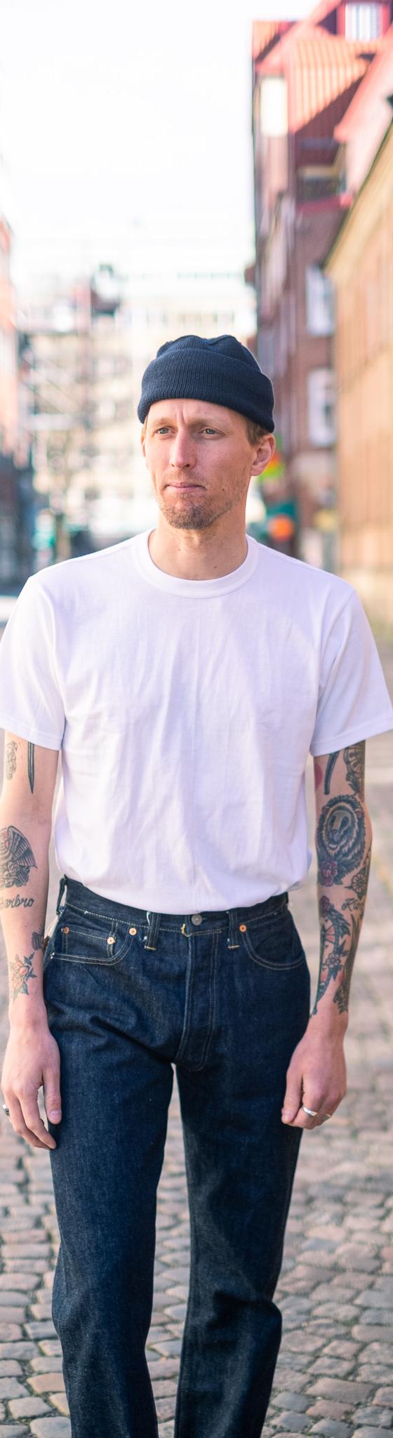 A man wearing a white Whitesville loopwheel pocket t-shirt with raw denim jeans and a black beanie on a cobblestone street