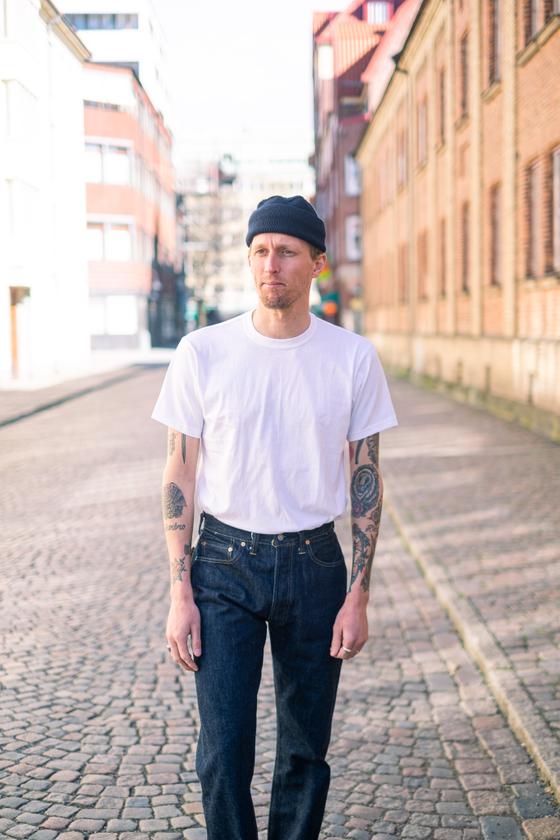 A man wearing a white Whitesville loopwheel pocket t-shirt with raw denim jeans and a black beanie on a cobblestone street