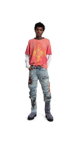 A man wearing a coral sun-print tee layered over a white long-sleeve with painted distressed Who Decides War jeans and boots