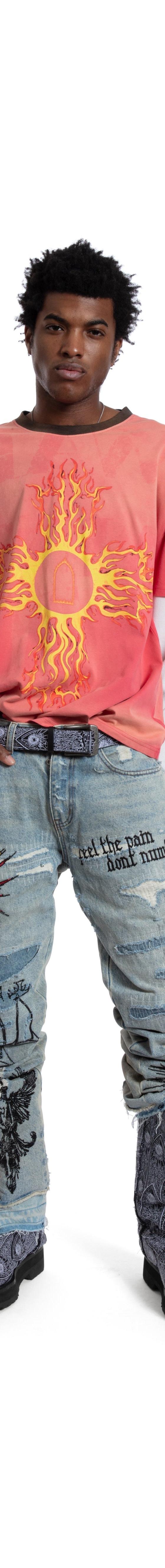 A man wearing a coral sun-print tee layered over a white long-sleeve with painted distressed Who Decides War jeans and boots